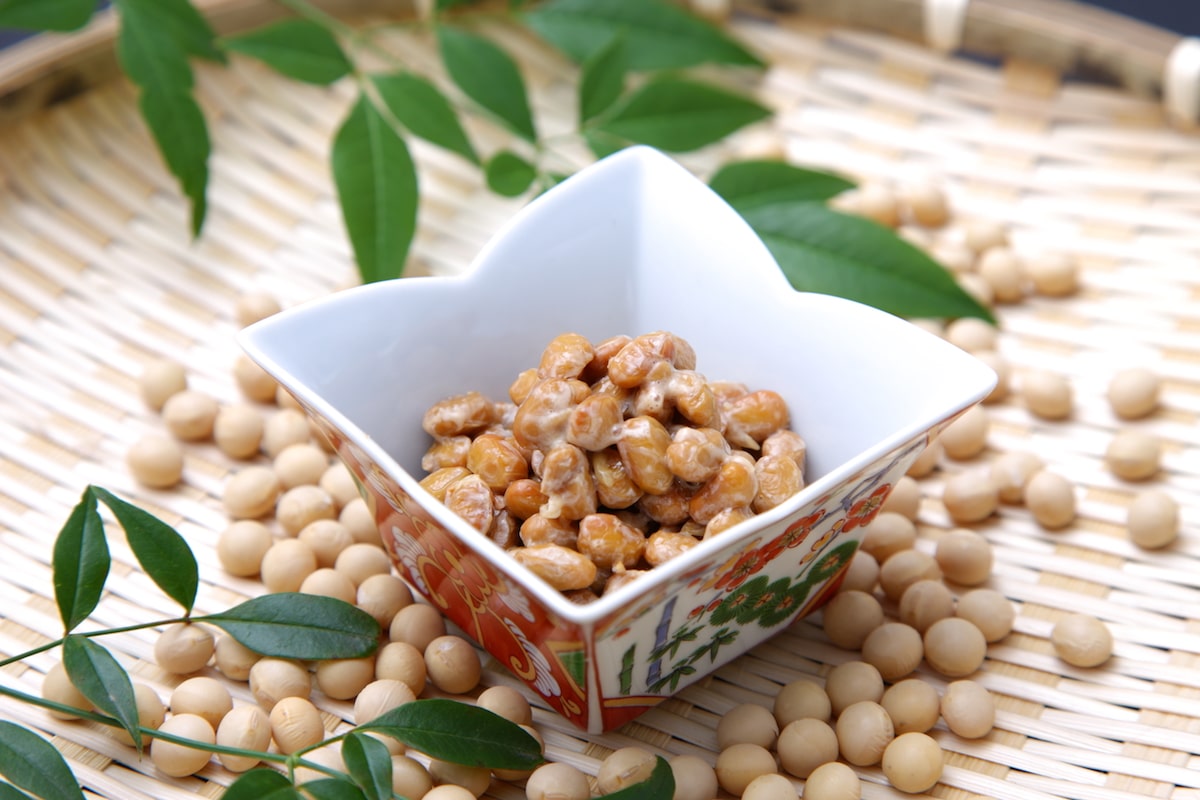 Scoop of fermented natto in a white bowl with raw soybeans sprinkled around